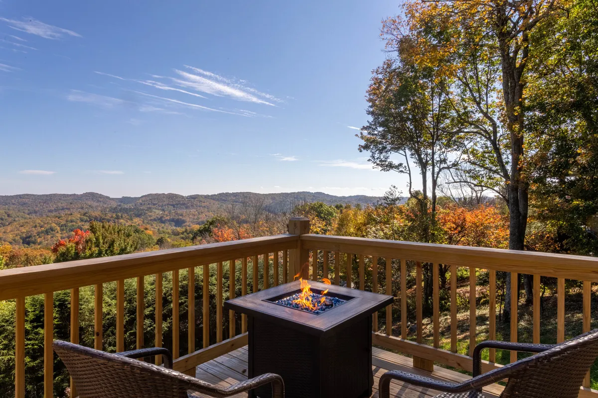 A warmly lit fire pit area sits on a deck, surrounded by comfortable seating and a classic deck railing, overlooking a breathtaking mountainous landscape. The cozy mood is perfect for sharing fireside moments or stargazing as the day winds down.
