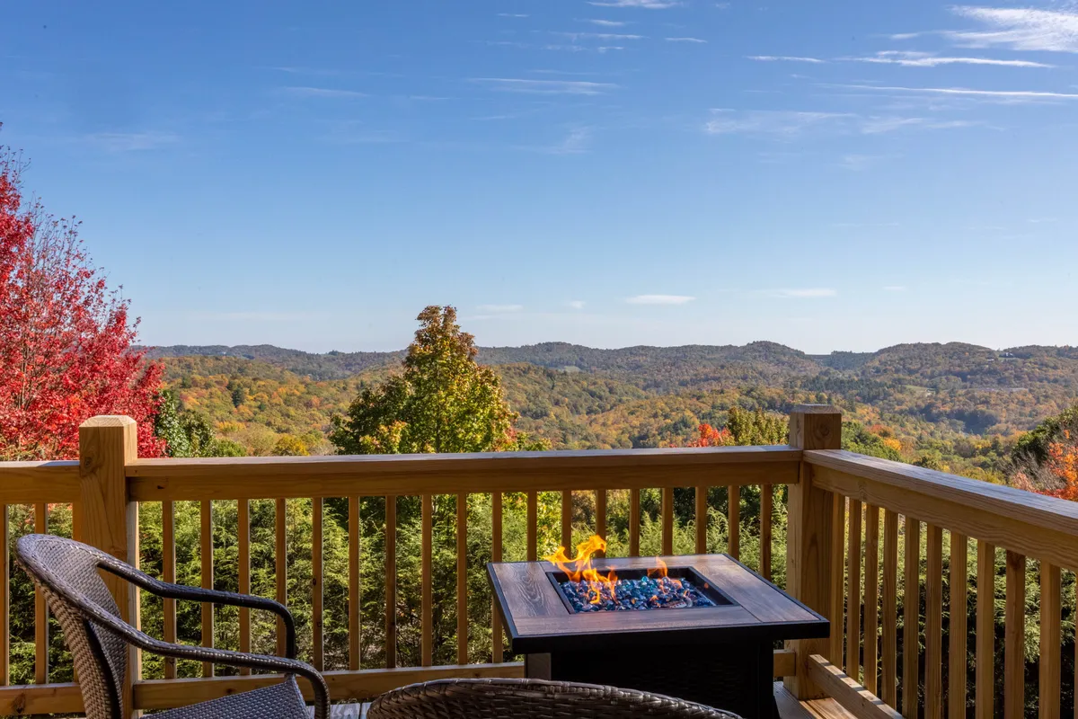 A cozy fire pit area on a wooden deck, framed by mountain views and autumn foliage. The scene evokes warmth and intimacy, ideal for evening relaxation or enjoying a quiet moment as the sun sets over the mountains.