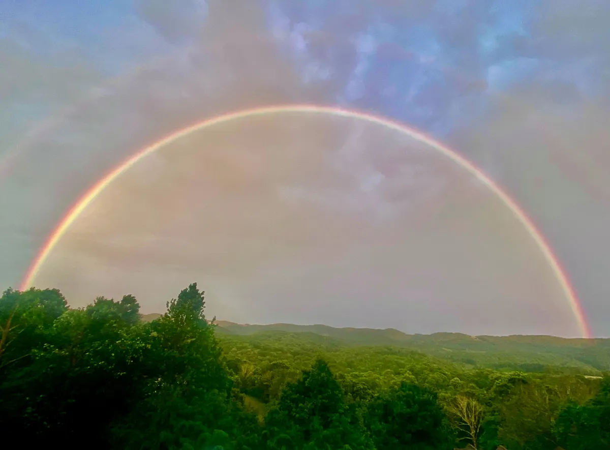 A stunning mountain vista is crowned by a brilliant rainbow arching over lush greenery. The image evokes a sense of wonder and tranquility, capturing the changeable beauty of the seasons and the magical moments nature can provide.