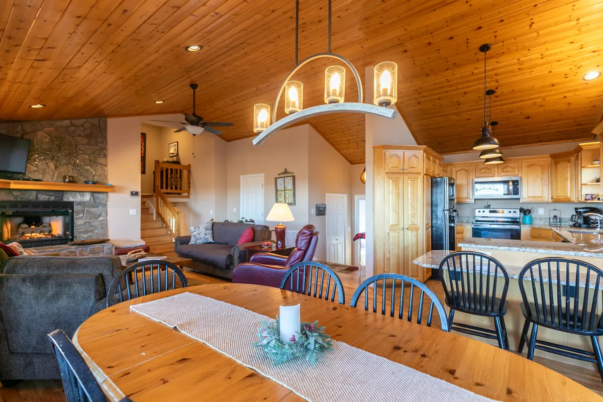 This image captures an inviting, open-concept main level, featuring a dining table set for gatherings, a cozy stone fireplace, a comfortable sofa area, and a view into the well-equipped kitchen. Natural light streams in, highlighting the spaciousness and warmth of the communal space.