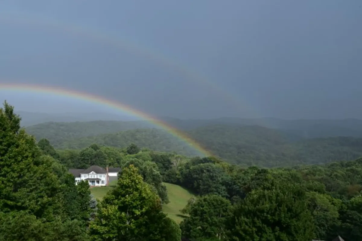 A picturesque view of the house against a backdrop of mountains, a rainbow, and lush greenery, capturing the broader setting and access to nature trails.