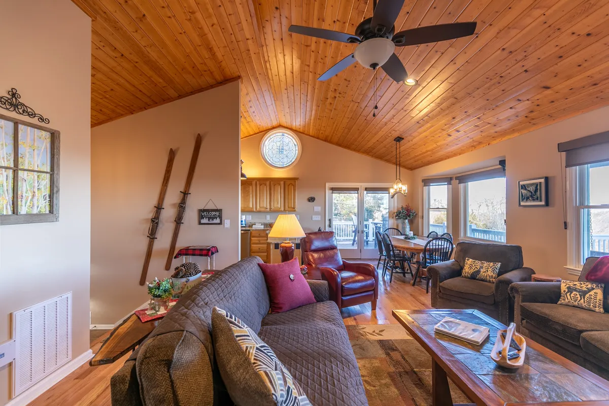 A wide-angle view of the main gathering area featuring a wooden ceiling, a large window drawing in sunlight and mountain views, plush seating, and a dining area set for family meals or laughter-filled evenings.