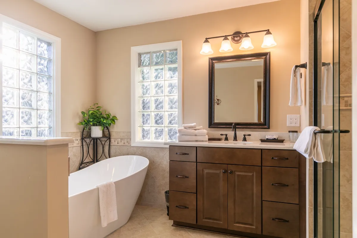 A spa-inspired bathroom with a freestanding bathtub, a dark wood vanity, large windows with glass blocks letting in soft natural light, and a mirror with a decorative frame. The setting is tranquil and luxurious, ideal for self-care and relaxation.