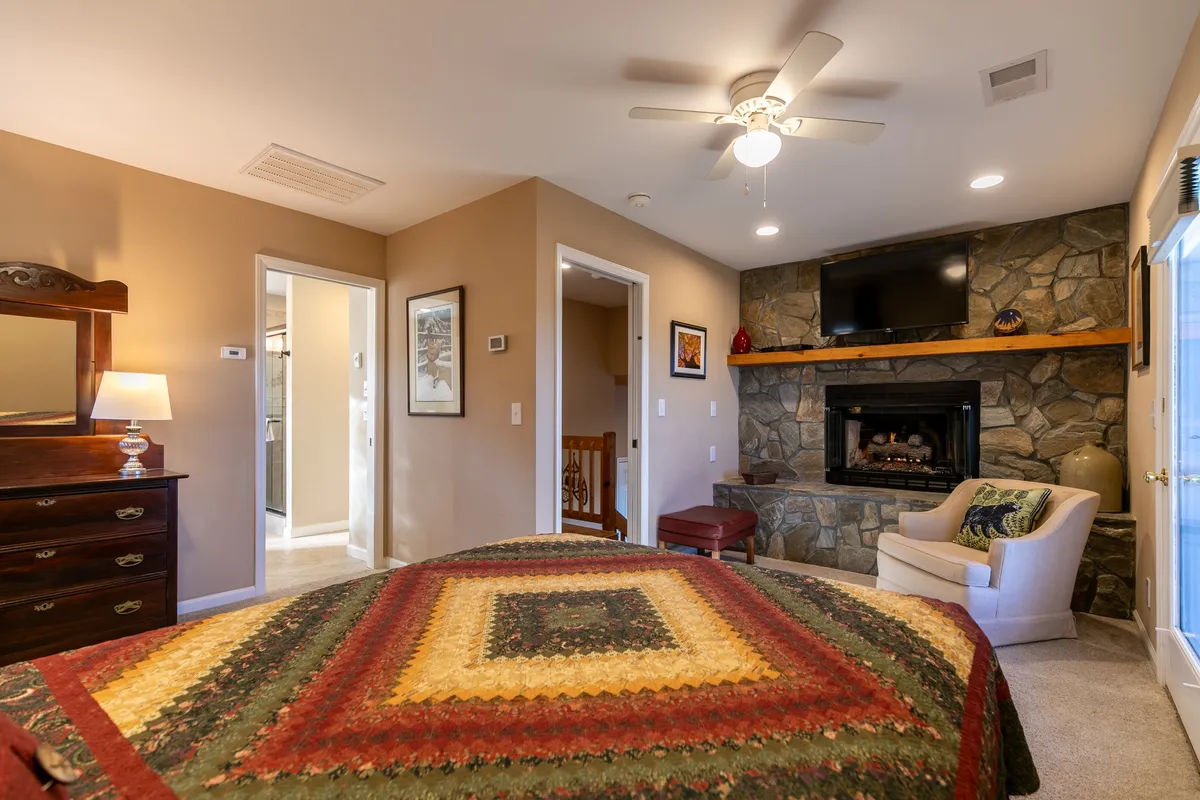 A cozy master bedroom featuring a bed with a colorful quilt, a charming stone fireplace beside the bed, an inviting armchair, and a wall-mounted TV. Soft lighting and rustic décor evoke warmth and comfort. The window by the fireplace hints at a view outside, possibly the Juliet balcony mentioned in the text.