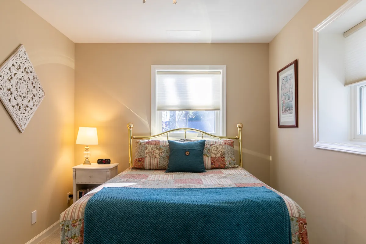 A bright and inviting bedroom featuring a bed with a decorative quilt, nightstand with lamp, framed artwork above the bed, and a window with blinds allowing natural light to filter in. The mood is peaceful and cozy, perfect for quiet moments.